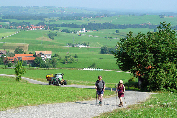 Zwei Wanderer gehen mit Wanderstöcken auf einem Wirtschaftsweg bergauf. Im Hintergrund fährt auf dem gleichen Weg ein Traktor mit Anhänger.