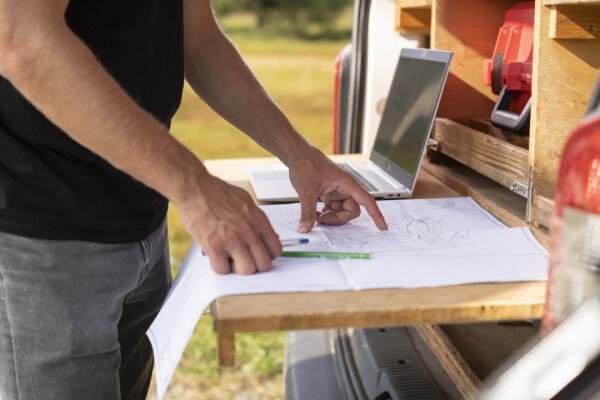 Handwerker an der Heckklappe seines Transporters mit ausgeklappter Arbeitsplatte auf der ein Notebook und eine Bauzeichnung liegt.