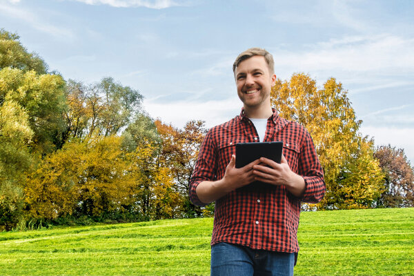Mann mit Tablet auf grüner Wiese, im Hintergrund sind Laubbäume.