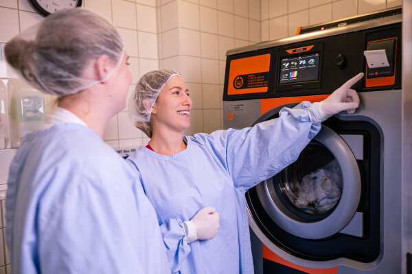 Zwei Hauswirtschafterinnen stehen in Arbeitskleidung an einer industriellen Waschmaschine.