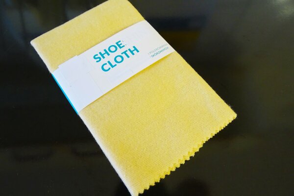 Ein gefaltetes, neues Mikrofasertuch vor einem dunklen Hintergrund, um das Tuch ist eine Banderole gebunden, auf der "Shoe Cloth" steht.