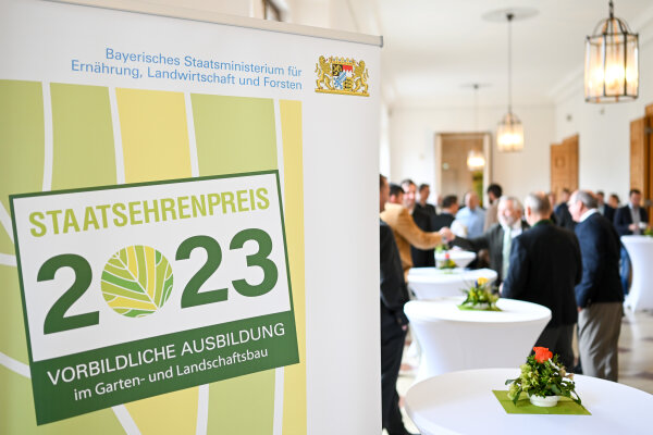 Blich in einen Saal mit Stehtischen und Personen. Links steht ein Banner mit Staatsehrenpreis 2023 vorbildliche Ausbildung.
