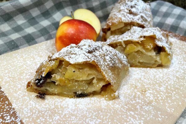 Ein Apfel-Zimt-Strudel auf einer Platte angerichtet mit einem geschnittenem Apfel und Puderzucker