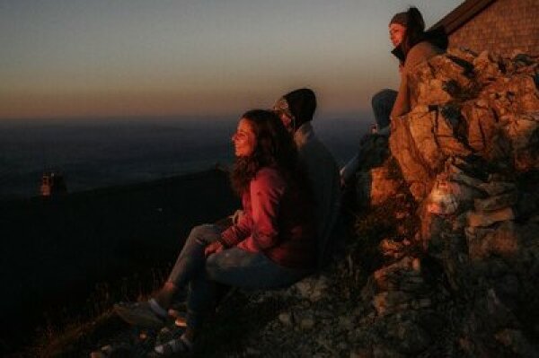 Drei junge Leuten sitzen auf einem Felsen in den Bergen und betrachten den Sonnenuntergang.