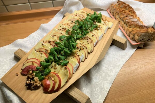 Auf einem Vesperbrett sind Apfel- und Camembertscheiben mit Honig und Feldsalat angerichtet