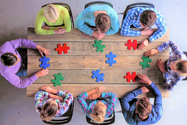 Acht Personen sitzen um einen Tisch, auf dem verschiedenfarbige, große Puzzleteile liegen.