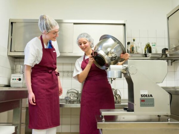 Eine Hauswirtschafterin leitet eine Auszubildende in einer Küche an.