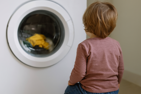 Man sieht ein den Rücken eines Kleinkindes, das in eine laufende Waschmaschine schaut.