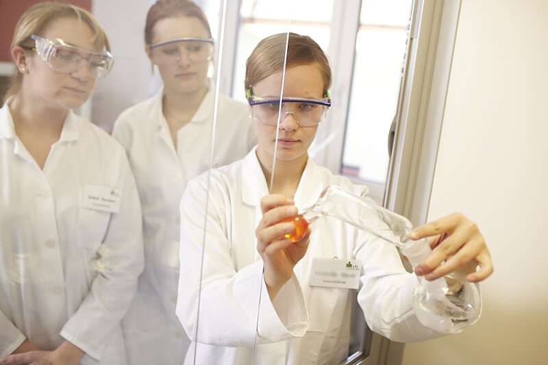 Eine Laborantin in weißem Kittel mit Schutzbrille hantiert mit Flüssigkeiten in Glasbehältern. Zwei andere Laborantinnen schauen zu.
