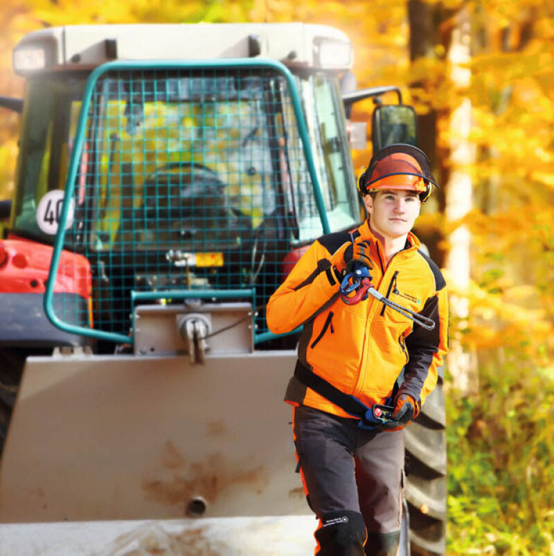 Ein Mann mit orangefarbenem Helm und Schutzkleidung vor einem Traktor im Wald