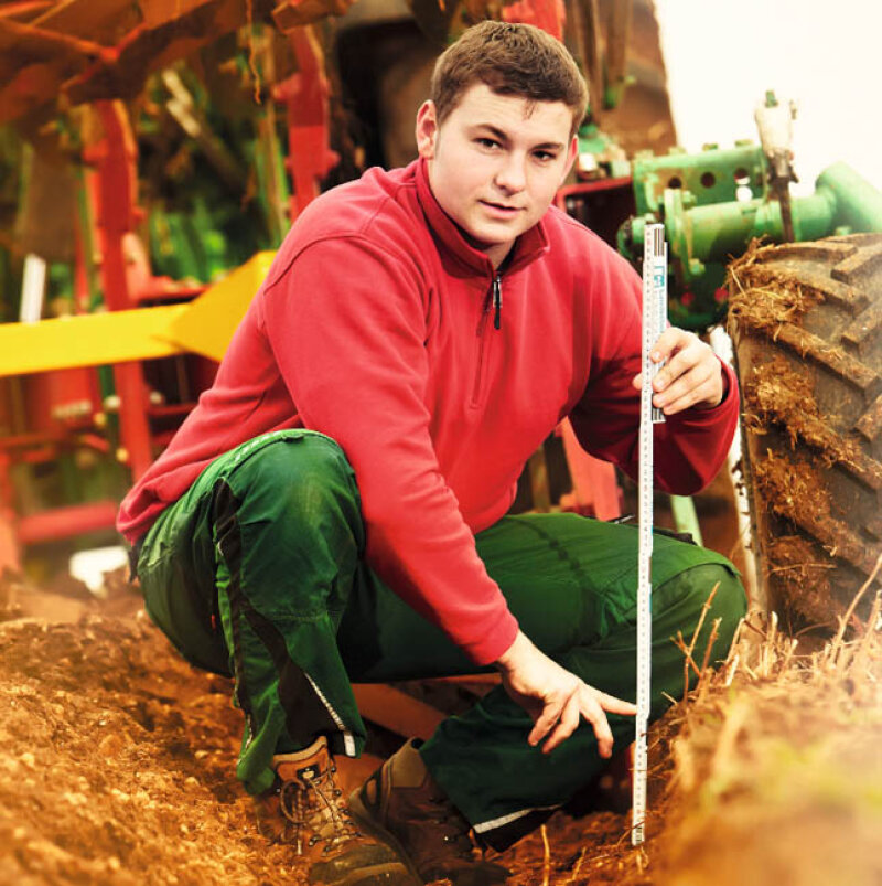 ein junger Mann in Arbeitskleidung kniet mit einem Meterstab vor einem Traktor