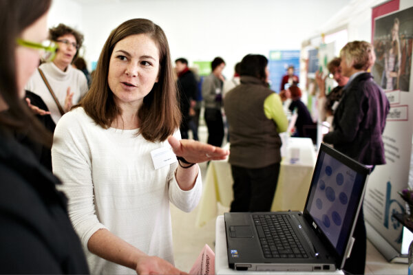 Frau steht neben Notebook an einem Stand und spricht zu Person