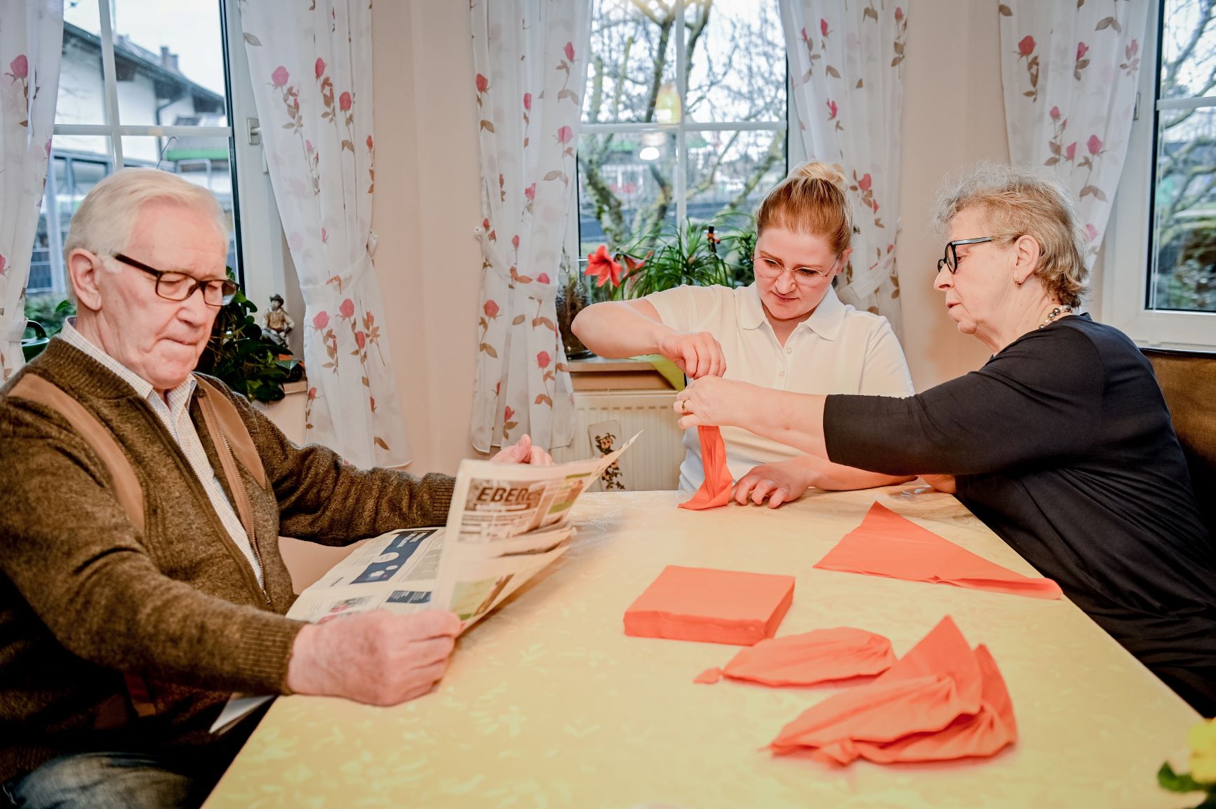 Seniorin und Pflegerin falten gemeinsam Servietten, Senior liest am Tisch Zeitung