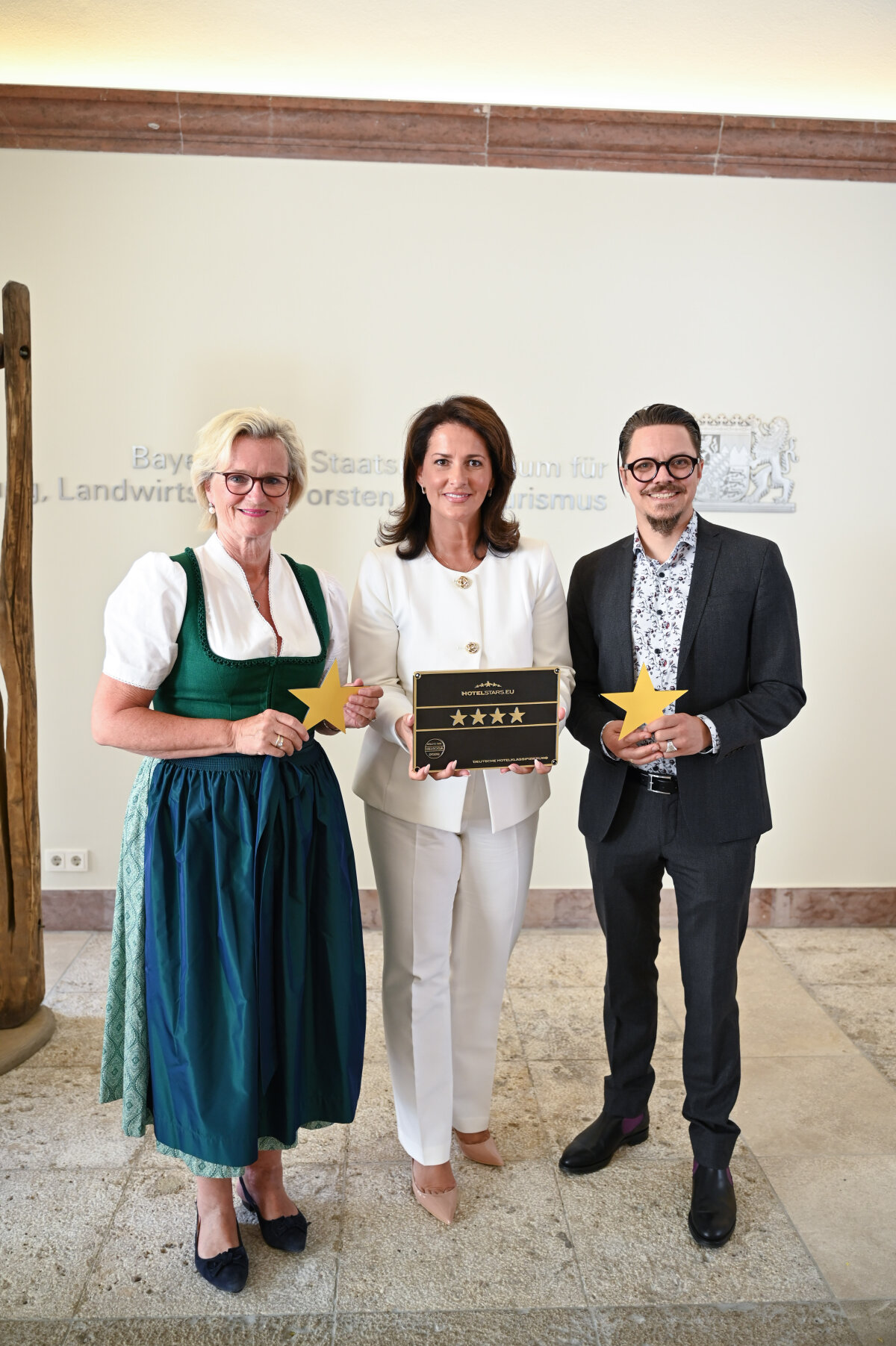 Das Bild zeigt Ministerin Kaniber, Angela Inselkammer sowie Wolfgang Wagner nebeneinander stehend im Tourismusministerium. Sie halten symbolische Sterne sowie eine Sterneklassifizierung mit vier Sternen in der Hand.