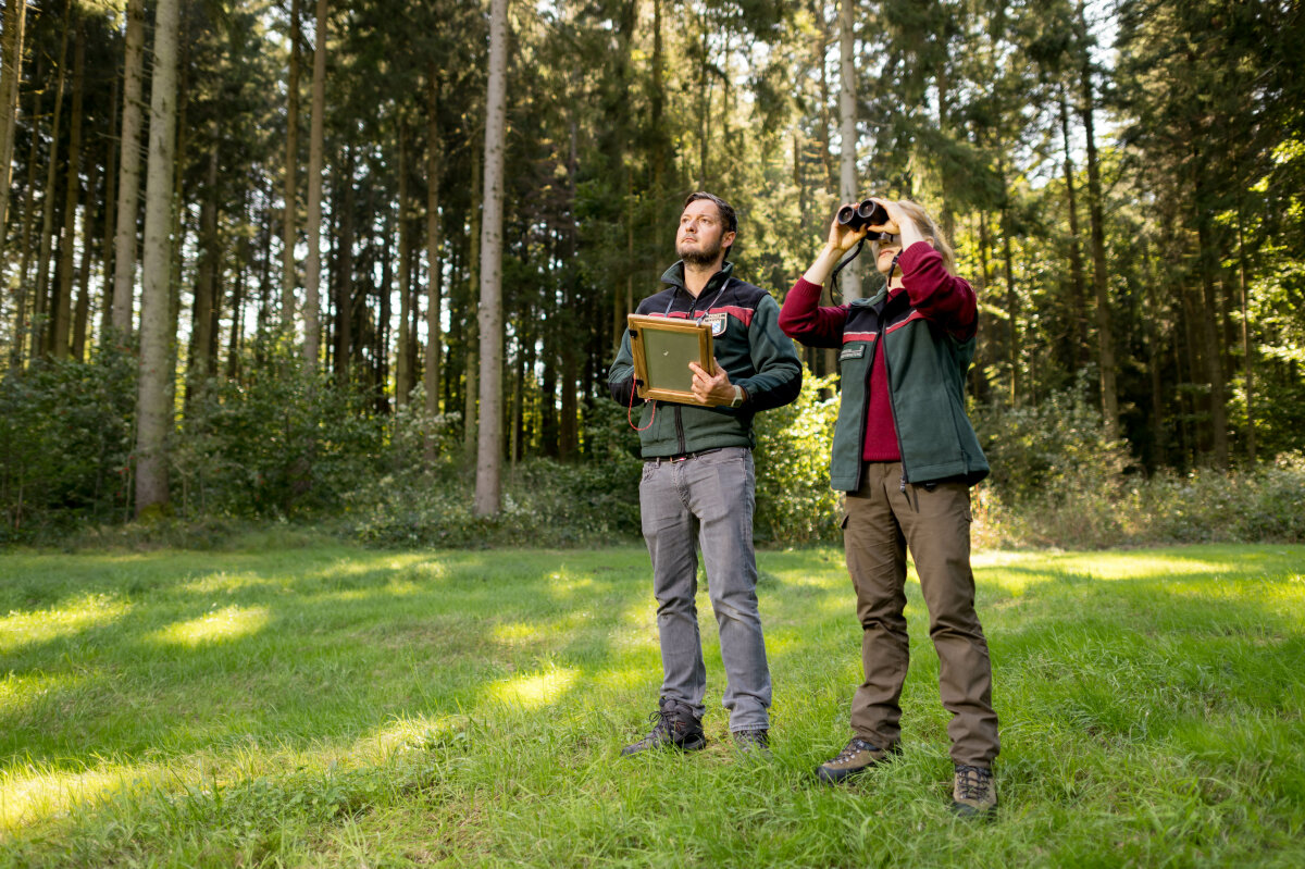 Das Bild zeigt zwei Förster in einem Waldstück mit Fernglas und Schreibutensilien in den Händen.