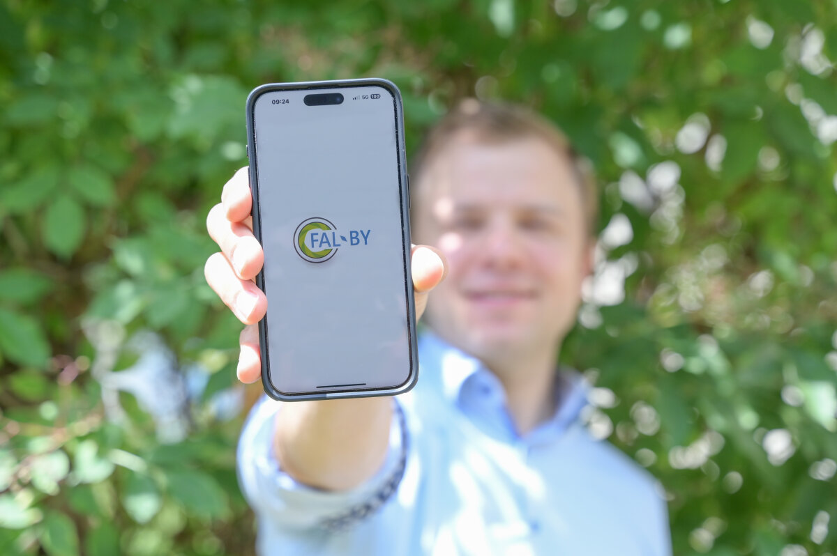 Das Bild zeigt einen Mann, der ein Handy in die Kamera hält, auf dem die Applikation FAL-BY zu sehen ist. Im Hintergrunde kann man grüne Blätter sehen. 
