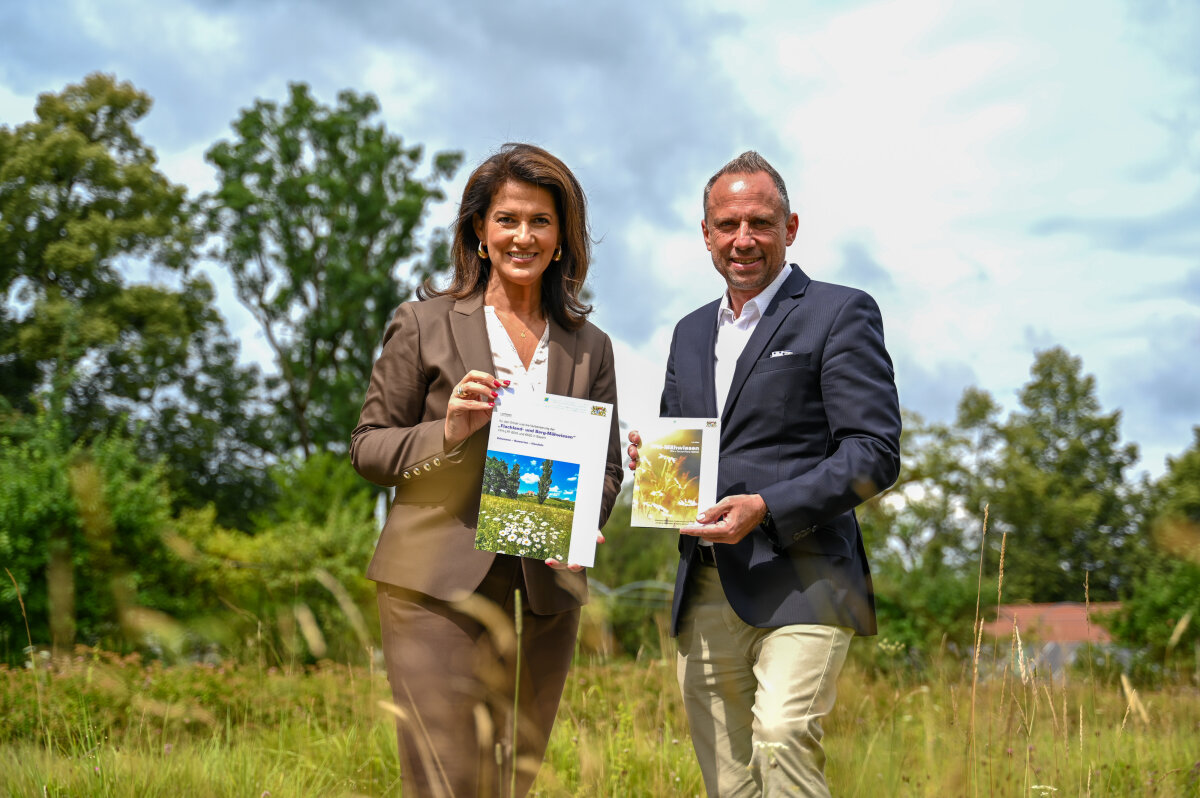 Ministerin Michaela Kaniber und Minister Thorsten Glauber stehen auf einer Wiese und halten Informationsbroschüren zu FFH-Mähwiesen in den Händen. Im Hintergrund sind Bäume und ein teils bewölkter Himmel zu sehen.