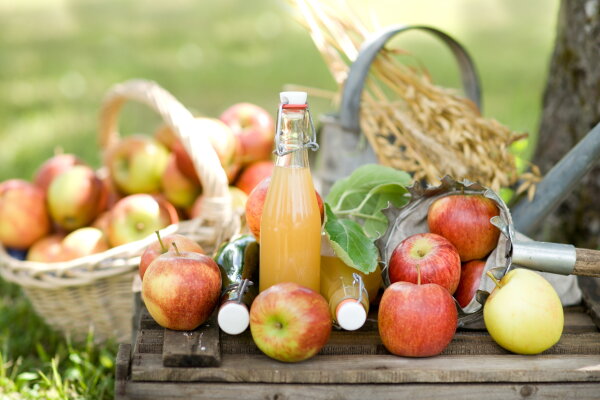 Korb mit Äpfeln steht auf einer Wiese. Auf einer Kiste liegen weitere Äpfel und Apfelsaftflaschen
