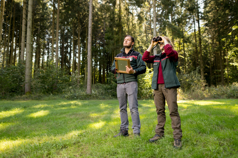 Förster und Försterin in einem Waldstück mit Fernglas und Schreibutensilien in den Händen