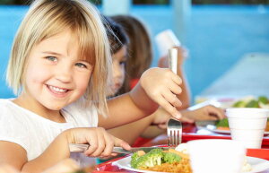 Kleines Mädchen mit Besteck in der Hand vor einem vollen Teller lacht in die Kamera