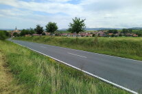 Eine leere Straße mit begrünten Randflächen und einem Dorf im Hintergrund.