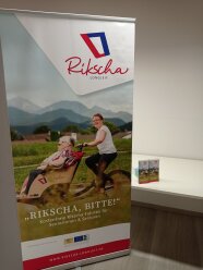 Werbeaufsteller mit Text Rkscha, bitte und Bild einer jungen Frau die zwei Seniorinnen auf einer Rikscha fährt.