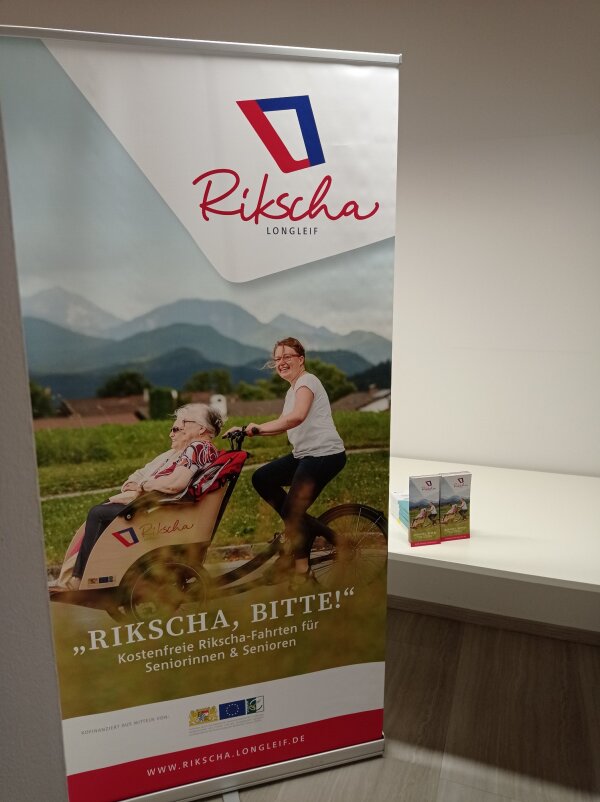 Werbeaufsteller mit Text "Rikscha, bitte!" und Bild einer jungen Frau die zwei Seniorinnen auf einer Rikscha fährt.