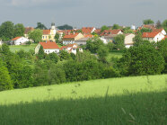 Im Vordergrund Wiese dahinter Baumreihe. Im Hintergrund ein Dorf mit ausschließlich roten Dächern. In hügeliger grüner Landschaft. 