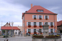 Interkommunales Bürgerzentrum Hofheim - 3-geschossiges Haus mit Walmdach, Sprossenfenstern, Fensterläden; links weitere Bebauung,; im Vordergrund Freifläche mit Brunnen