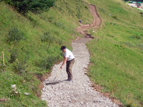 1,5 m Meter breiter Alpweg, auf dem ein Arbeiter den eingebrachten Schotter ebnet.