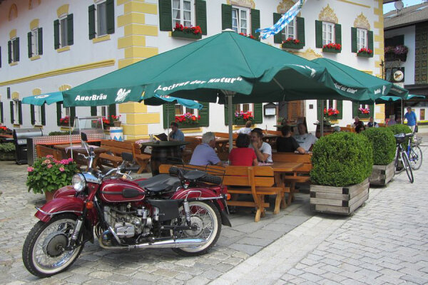 Ein Wirtshaus mit eine Tischgruppe vor dem Eingang, auf denen ein paar Menschen sitzen.