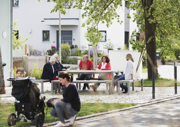 Auf einem Dorfplatz unterhalten sich mehrere Personen an einem öffentlichen Tisch mit Bänken. Im Vordergrund befindet sich eine Mutter mit Kinderwagen.