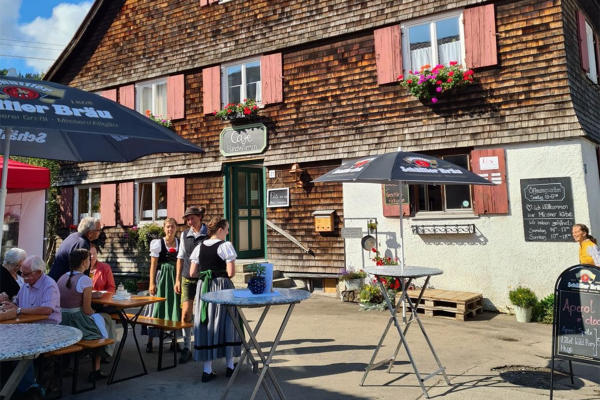 Vor einem historischen Gebäude mit Holzvertäfelung ist ein kleines Cafe aufgebaut. Mehrere Personen sitzen an Tischen.