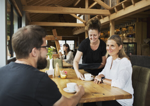 Eine Frau serviert einen Kaffee an einem Tisch. Am Tisch sitzen eine Frau und ein Mann, die sich unterhalten.