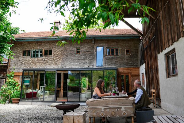 Ein Blockhaus ist mit einem zeitgemäßen Wintergarten ergänzt. Im Vordergrund sitzt in sommerlichen Ambiente eine Frau und ein Mann am Tisch.