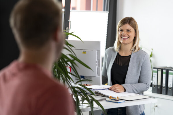 Eine Person unscharf im Vordergrund; im Hintergrund steht eine lächelnde junge Frau an einem Arbeitstisch