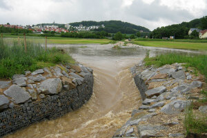 Angestautes Wasser an einer Bodenwelle, das durch den mit Gabionen befestigten Durchlass fließt.