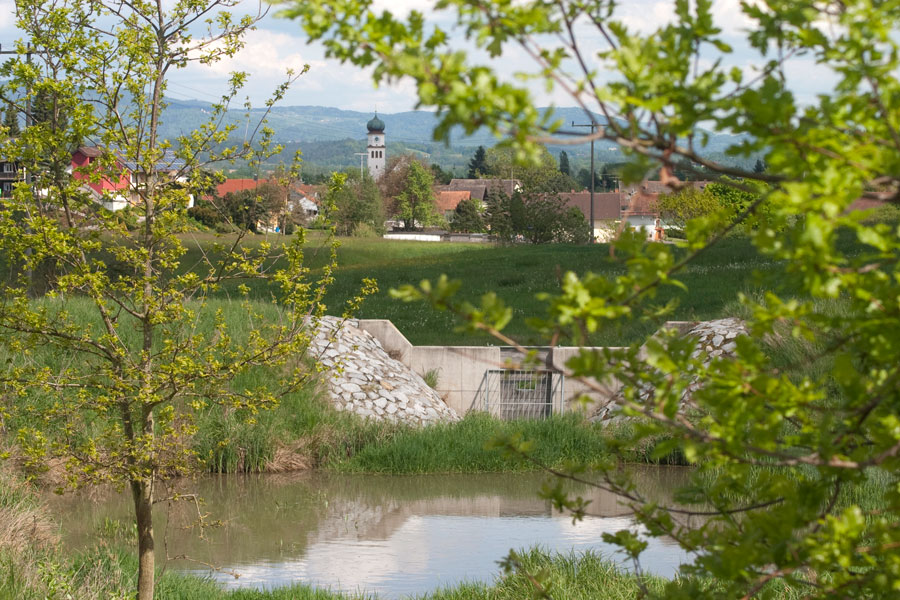 Wasserrückhaltebecken mit Stau- und Ablassbauwerk. Im Hintergrund ein Dorf.