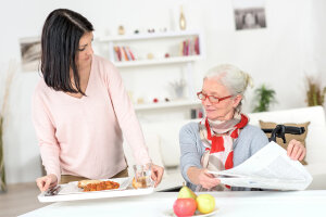 Frau stellt Tablet mit Essen und Getränk auf Tisch, an dem Seniorin sitzt