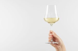 Eine Hand hält ein langstieliges Weinglas mit Weißwein vor hellem Hintergrund in die Höhe.