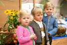 Drei kleine Kinder posieren für die Kamera. Im Hintergrund ist ein Korb mit Äpfeln und Blumen