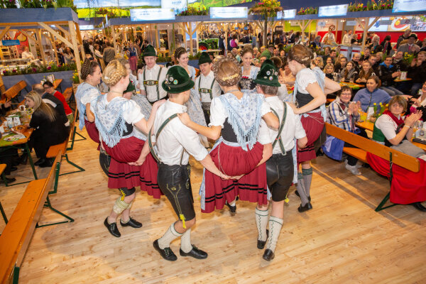 Männer in Lederhosen tanzen im Kreis und halten sich an den Händen fest. Frauen in Dirndel sitzen jeweils zwischen den Männern auf ihren Händen. Im Hintergrund sind Zuschauer.