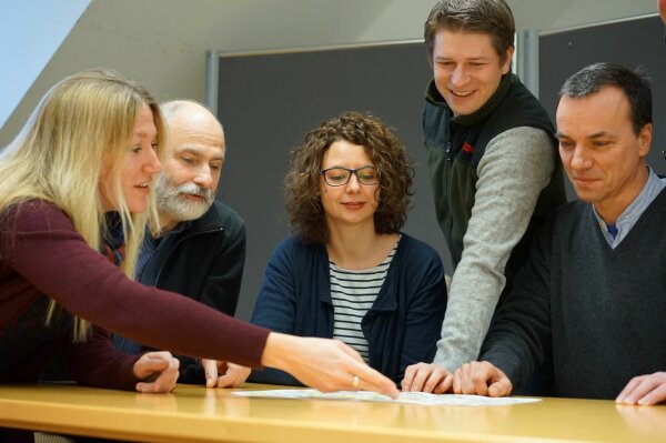 Zwei Frauen und drei Männer diskutieren in einem Besprechungsraum über einen auf dem Tisch liegenden Plan.