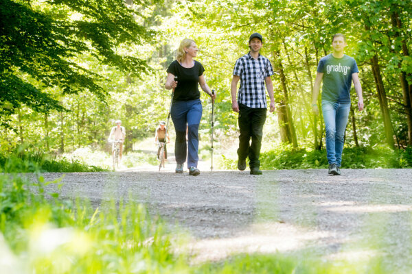 Drei Personen gehen im Wald spazieren. Im Hintergrund kommen zwei Radfahrer.