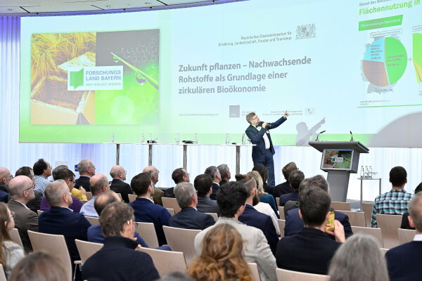 Mann mit Mikrofon auf der Bühne zeigt auf eine Präsentation, vor ihm das Publikum.