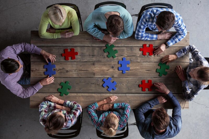 Acht Personen sitzen um einen Tisch, auf dem verschiedenfarbige, große Puzzleteile liegen