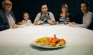 Ein Senior, ein Kind, ein Arbeiter, eine Schülerin und ein Student sitzen an einem Tisch und schauen mit erwartungsvollem Blick auf einen Kantinenteller mit einem leckeren Gericht.