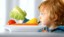 Junge sitzt vor einem Teller mit sorgfältig arrangiertem rohem Gemüse und hält ein Salatblatt in der Hand