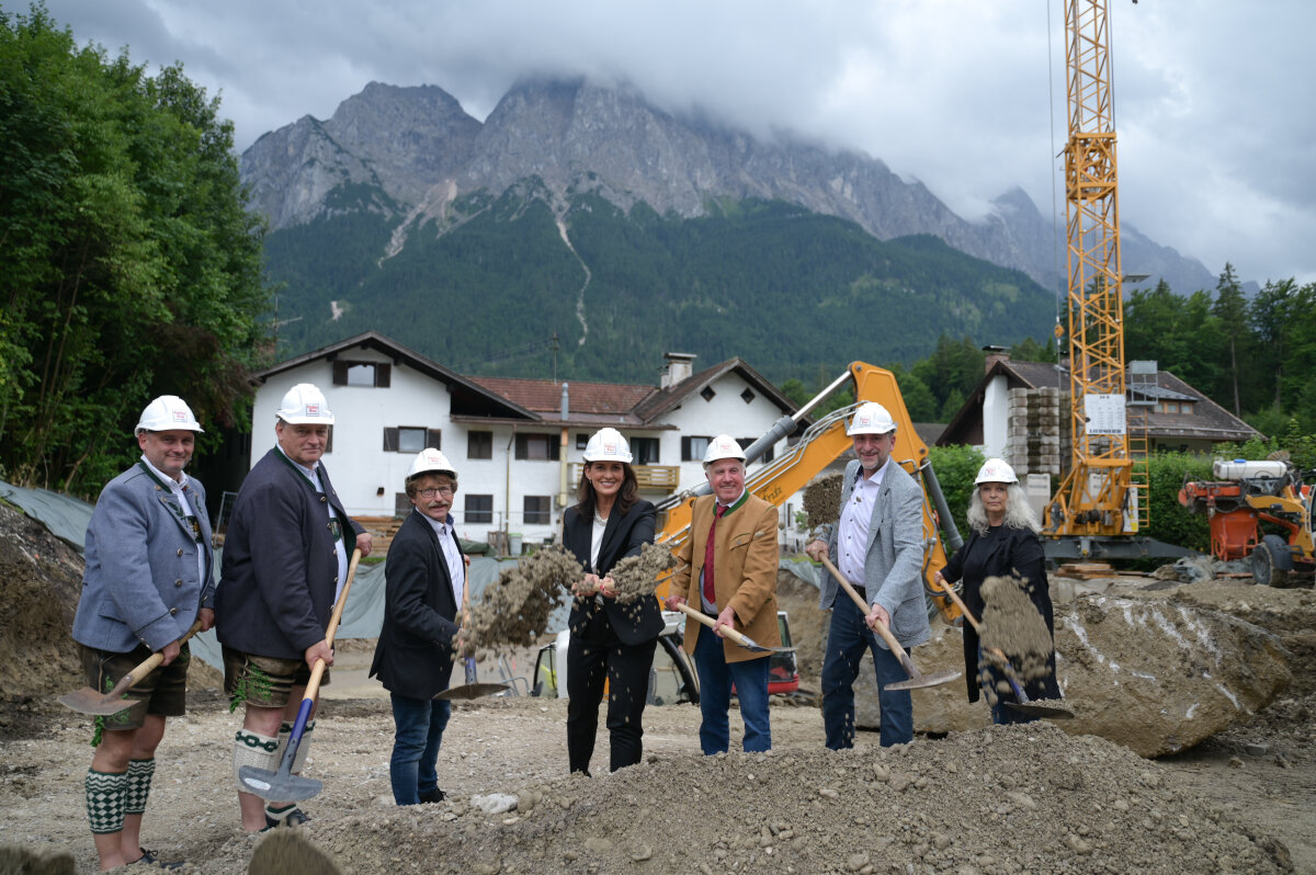 Ministerin Michaela Kaniber (Mitte) beim symbolischen Spatenstich mit sechs Mitwirkenden auf einer Baustelle in alpiner Umgebung. Alle tragen Bauhelme und halten Spaten. Im Hintergrund: Bagger, Baukran, Häuser und Berge.