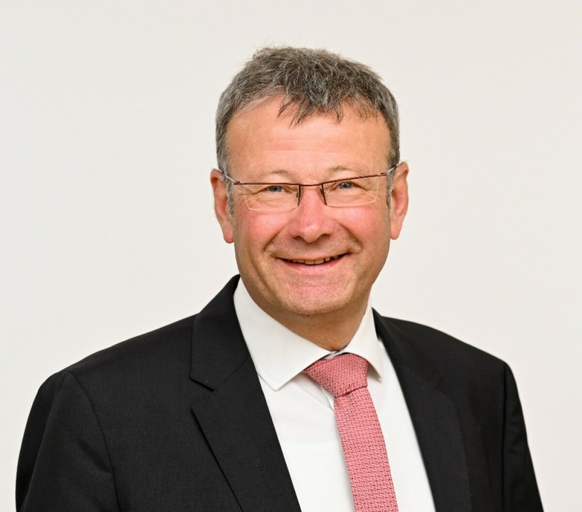 Dr. Becker lächelt vor einem hellen Hintergrund in die Kamera. Er trägt einen Anzug mit Krawatte.